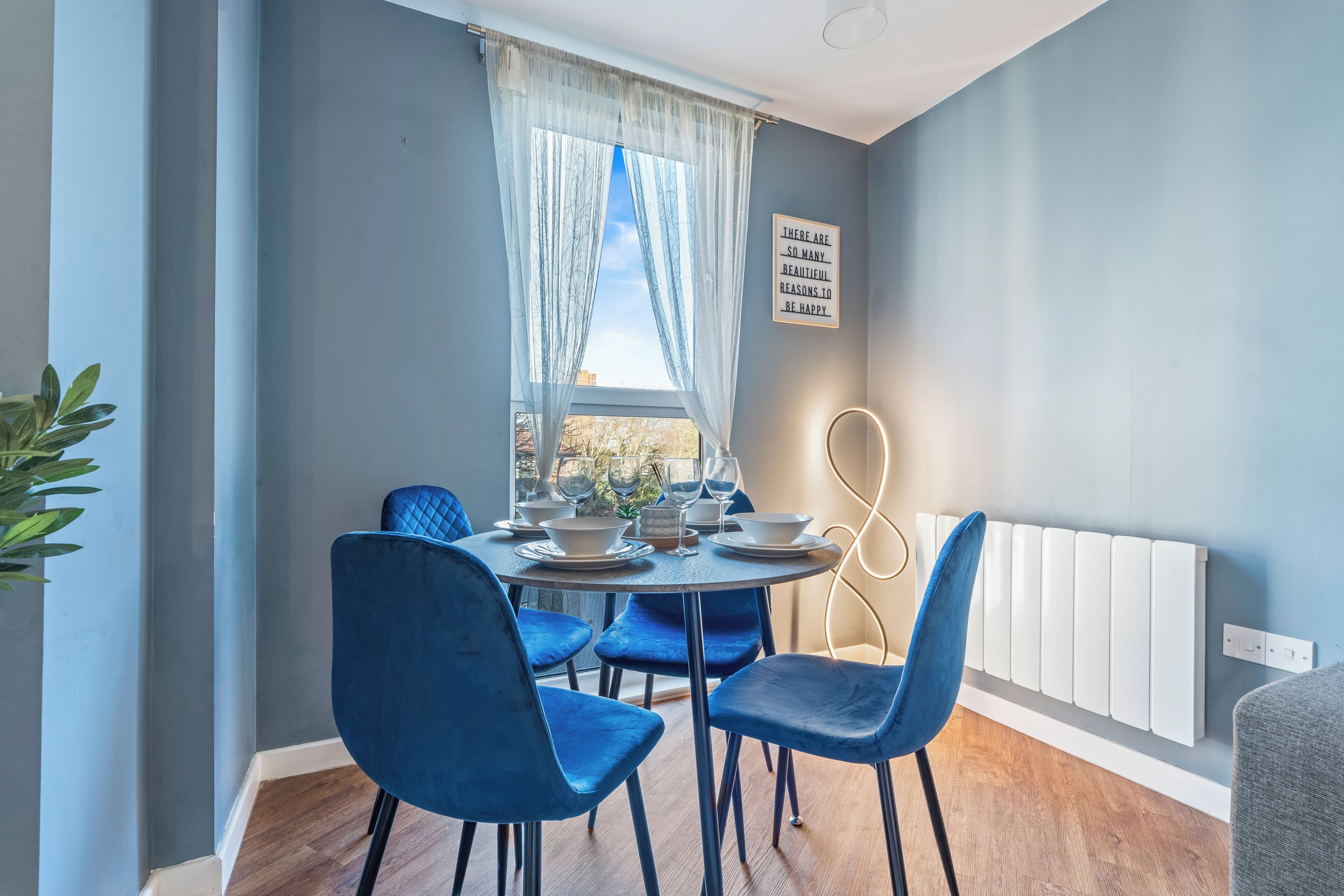 A dining area with a round table, blue chairs, and a window with sheer curtains.
