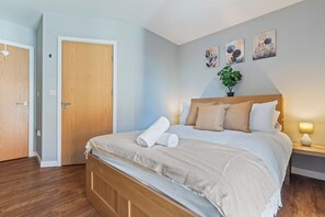 Ein ordentlich bezogenes Bett mit hölzernem Kopfteil, ein Nachttisch mit Lampe und eine Wand mit drei gerahmten Kunstwerken.