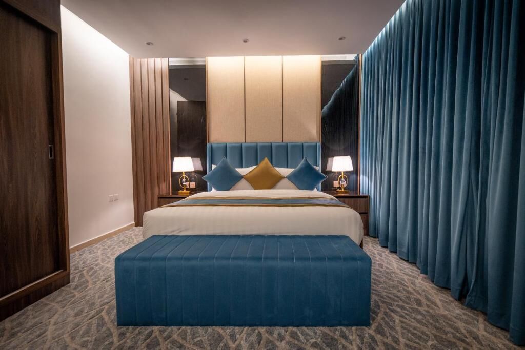 Habitación de hotel con una cama grande, una mesita de noche, un pouf azul y vistas de la ciudad a través de la ventana.