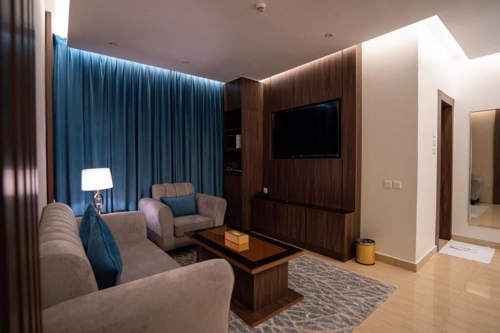 Una sala de estar moderna con un sofá, sillones, una mesa de centro, un televisor de pantalla plana montado en una pared de madera y un gran ventanal con cortinas azules.