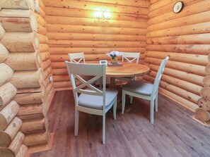 A wooden cabin interior with a round table and four chairs, a wall clock, and a small vase with flowers on the table. - The Log Cabin (Ormskirk)