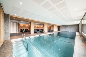 Une piscine intérieure moderne avec de l’eau cristalline, une terrasse en bois et des chaises longues.