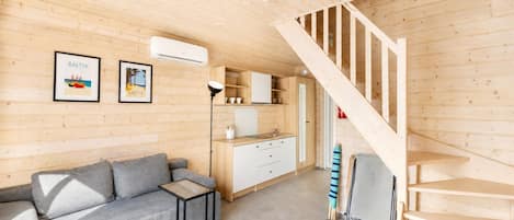 A compact room with a wooden ceiling, a grey sofa, a small table, a kitchen area with cabinets, and a wooden staircase.