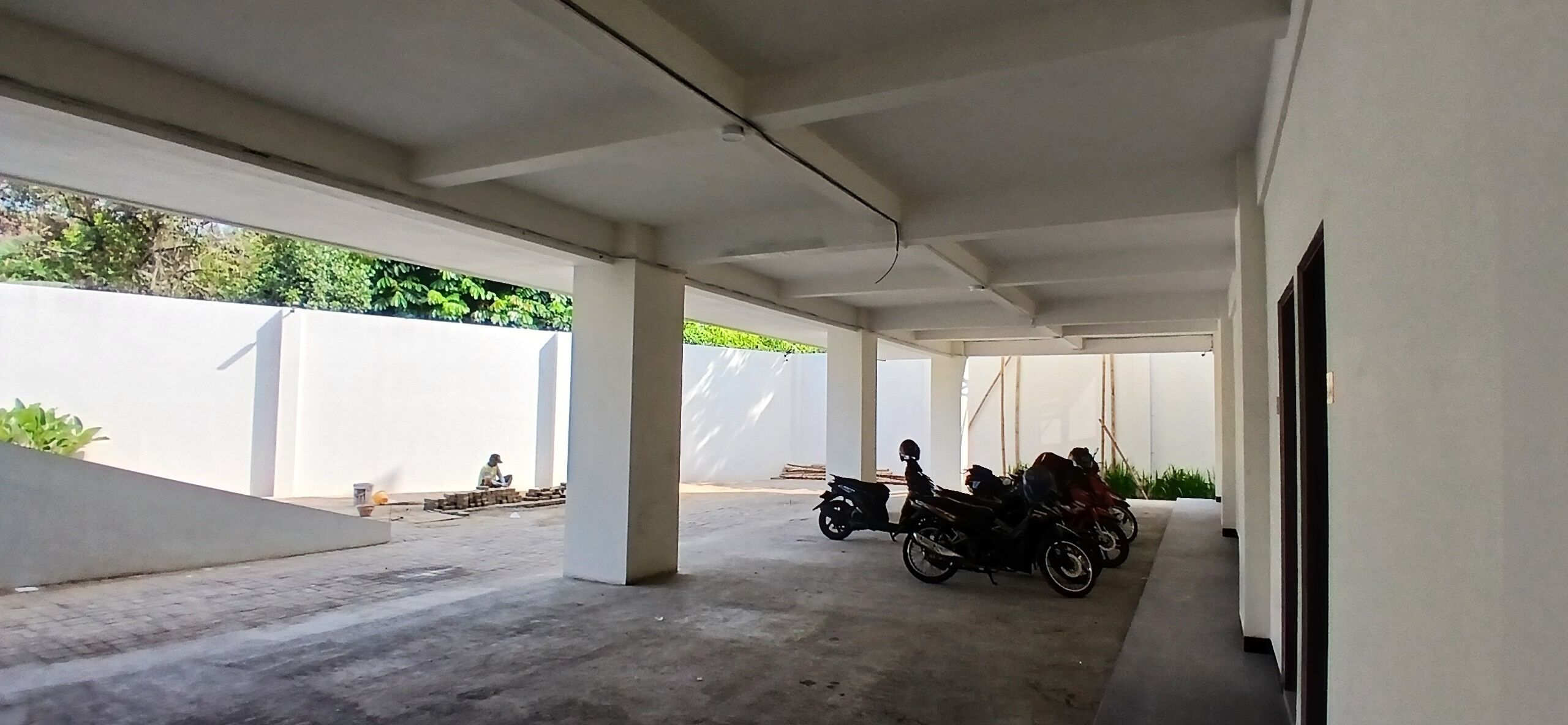 A covered outdoor area with concrete flooring, white walls, and a row of parked motorcycles.