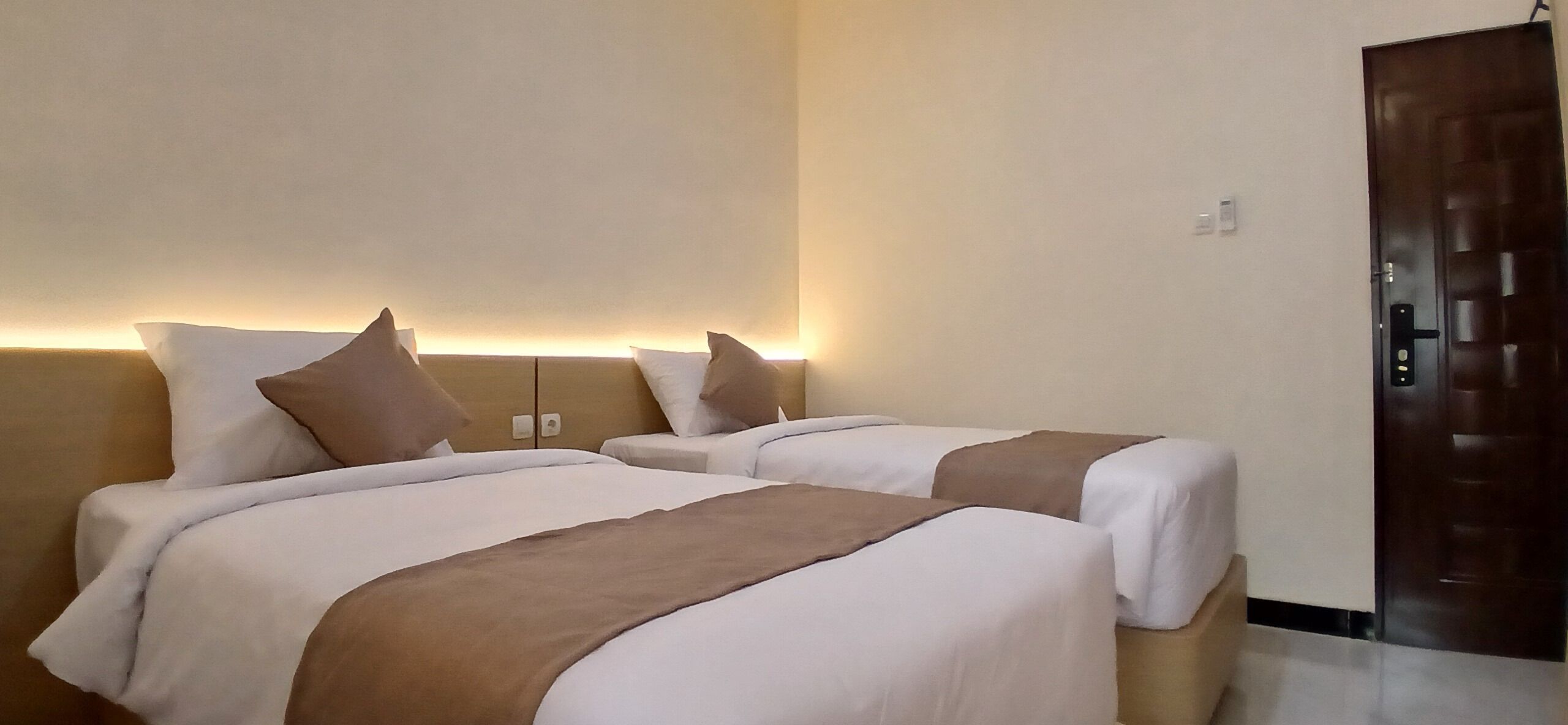 A hotel room with two beds, each with white and brown bedding, a wooden door, and a wall-mounted light fixture.