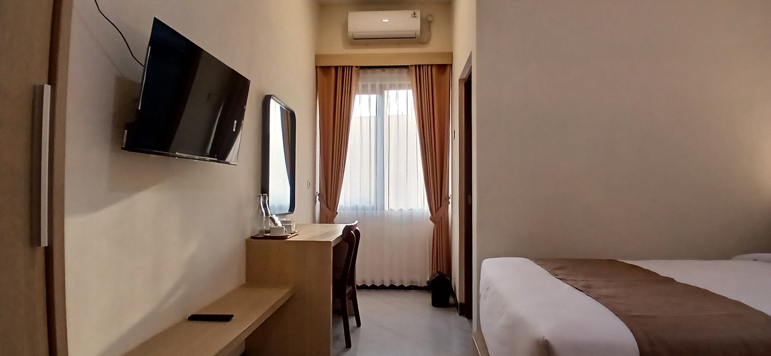 Habitación de hotel con una cama, un escritorio con silla, un televisor montado en la pared y una ventana con cortinas.