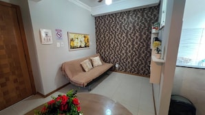 Ein Zimmer mit einer Couch, einem Wandradiator, einem gerahmten Blumenbild, einer Topfpflanze und einem Fliesenboden.
