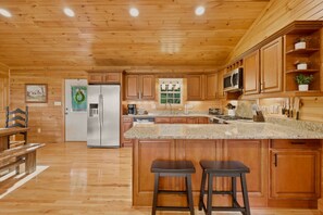 A spacious kitchen with wooden cabinets, a stainless steel refrigerator, a granite countertop, and a large island with two stools. - New listing! Cabin with mountain view-fire pit-hot tub-privacy-8 min to ASU (Boone)