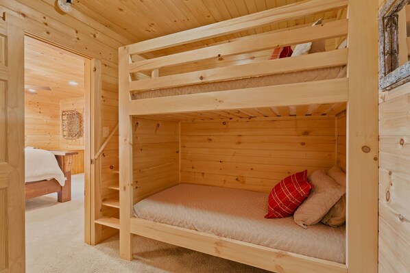 A wooden bunk bed with a mattress and pillows, a wooden table with a white cloth, and a woven basket on the wall.