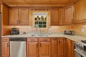 A kitchen with wooden cabinets, a stainless steel dishwasher, a built-in oven, a coffee maker, and a window with hanging lights.