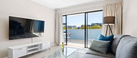 Una sala de estar moderna con un televisor de pantalla plana, una mesa de centro de vidrio, un sofá con cojines, una lámpara de pie y un ventanal corredizo que da a un balcón exterior.