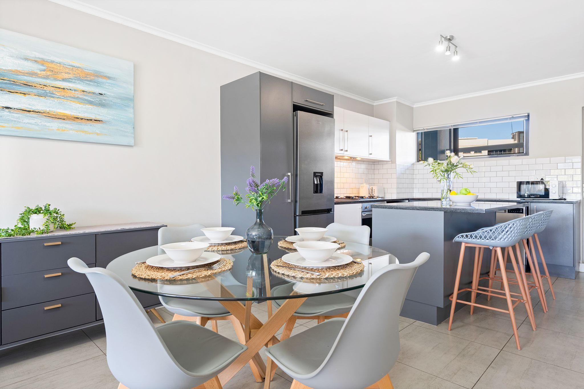 Cocina moderna con una isla central, zona de comedor con mesa de vidrio y barra con taburetes.