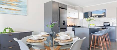 Cocina moderna con una isla central, zona de comedor con mesa de vidrio y barra con taburetes.