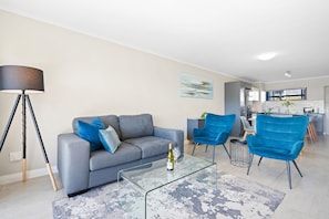 Ein modernes Wohnzimmer mit einem grauen Sofa, zwei blauen Sesseln, einem Glastisch und einer Stehlampe.