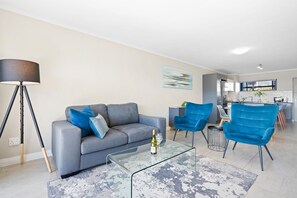 Uma sala de estar moderna com um sofá cinzento, duas poltronas azuis, uma mesa de centro de vidro e um abajur.