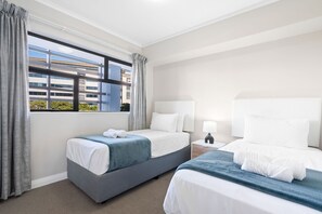 Quarto de hotel com duas camas, uma janela com cortinas, mesa de cabeceira com abajur e vista para um edifício.