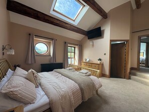 A bedroom with a large bed, a wooden headboard, a skylight, a TV mounted on the wall, and a dresser. - The Shippon (Ormskirk)
