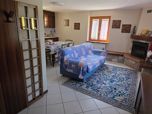 A living room with a sofa, dining table, chairs, a fireplace, and a window with curtains. - House with garden and garage near the slopes (San Pietro)