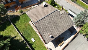 An aerial view of a residential building with a sloped roof, a fenced yard, and a street visible in the background. - House with garden and garage near the slopes (San Pietro)