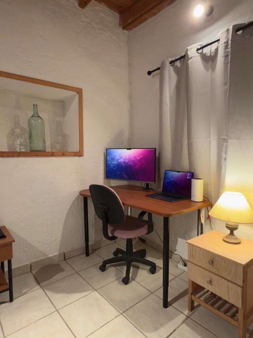 A room with a desk, two monitors, a laptop, a chair, a lamp, and a small cabinet.