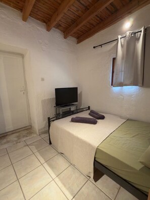 A small, single-bed room with a television on a stand, a window with curtains, and a tiled floor. - A stone retreat 40m from the sea, in a private 1,200m² estate on Aegina island. (Aegina)