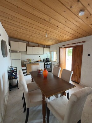 A dining area with a wooden table and chairs, a kitchen in the background, and a window with curtains. - A stone retreat 40m from the sea, in a private 1,200m² estate on Aegina island. (Aegina)