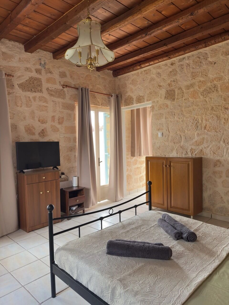 Une chambre avec un mur en pierre, un lit recouvert d’une couvre-lit à motifs, une armoire en bois, un téléviseur posé sur un meuble en bois et un lustre.