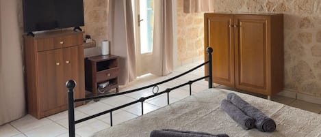 Un dormitorio con pared de piedra, una cama con cubrecama estampado, un armario de madera, un televisor sobre un mueble de madera y una lámpara de araña.