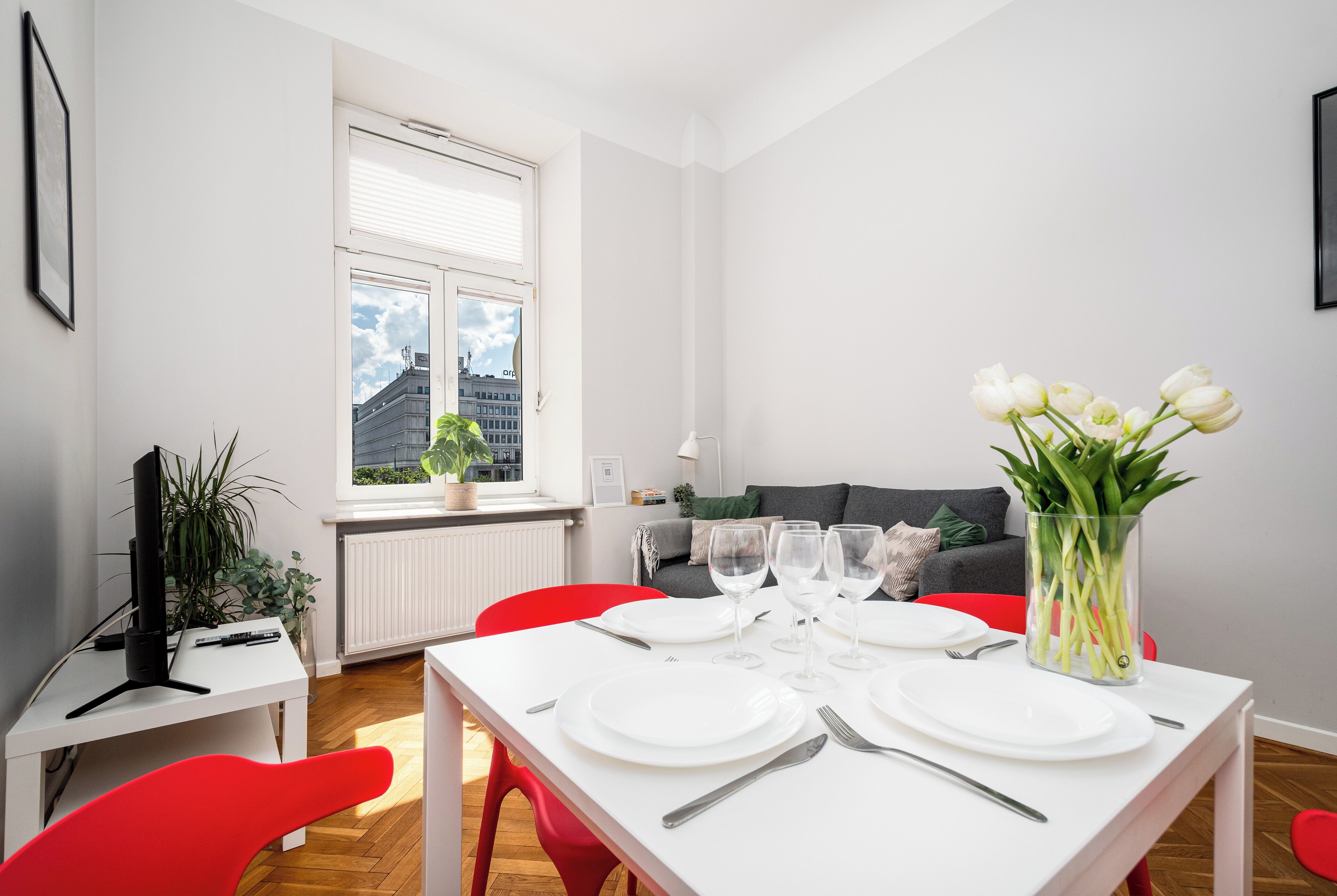 Un comedor con una mesa preparada para cuatro personas, un sofá al fondo y una ventana con persianas.