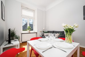Un comedor con una mesa preparada para cuatro personas, un sofá al fondo y una ventana con persianas.