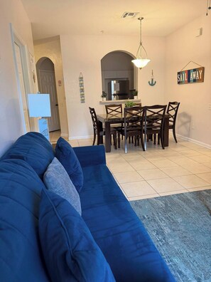 A living room with a blue sofa, a dining area with a table and chairs, a kitchen in the background, and a 'Sail Away' sign on the wall. - Windsor Palms apartment near Disney (Kissimmee)