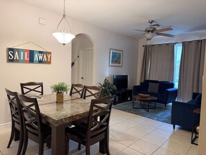 A dining area with a marble table and dark wooden chairs, a living room with a dark blue sofa and a television, and a ceiling fan. - Windsor Palms apartment near Disney (Kissimmee)
