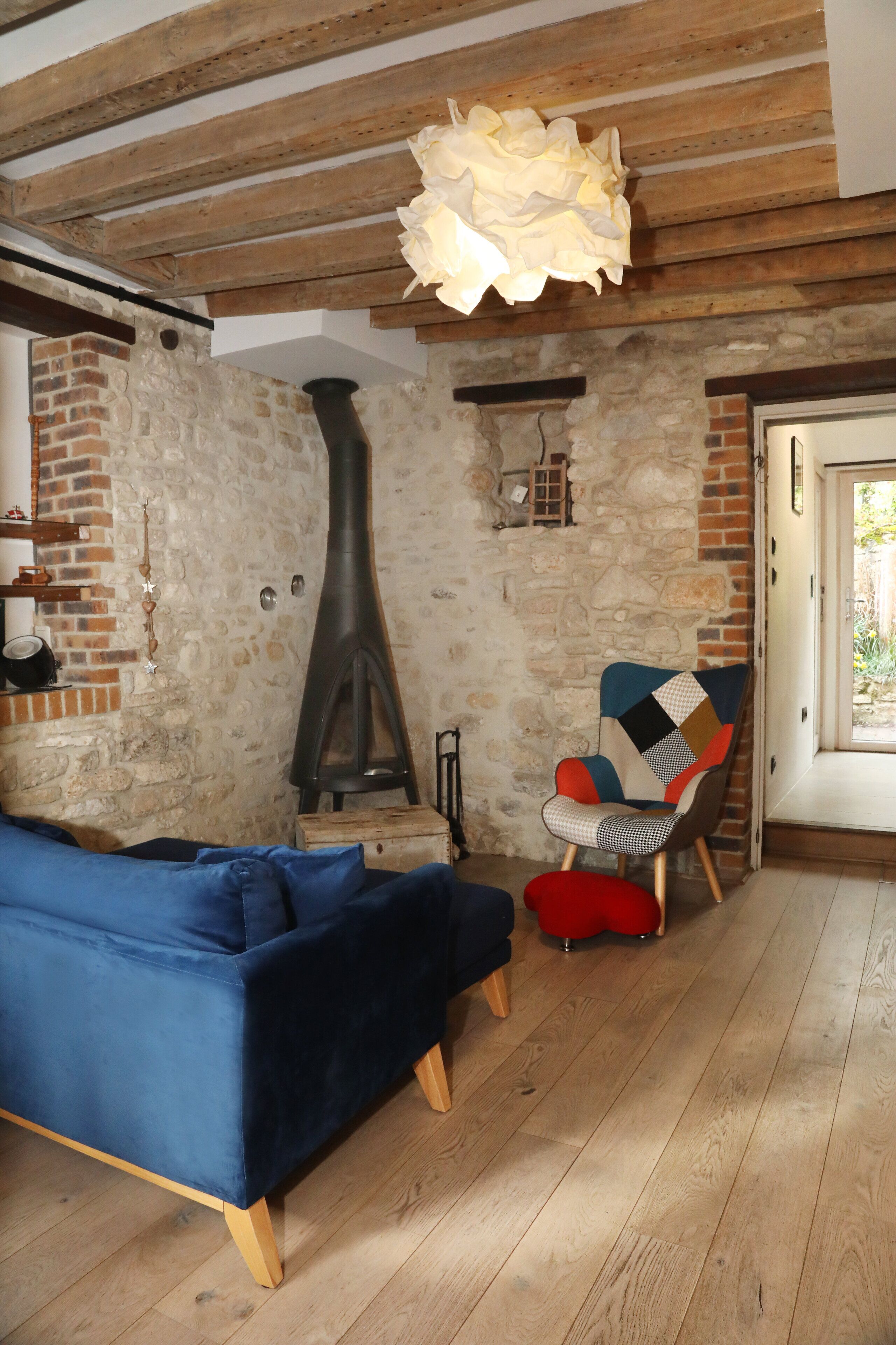 Une pièce rustique avec des poutres apparentes, un mur en pierre, un canapé bleu, un fauteuil rouge et une grande cheminée noire.