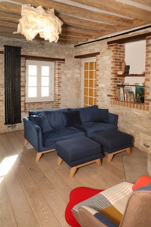 Ein Zimmer mit einem blauen Ecksofa, einem roten Sessel, Holzboden, einer Ziegelwand und einer hängenden Leuchte.
