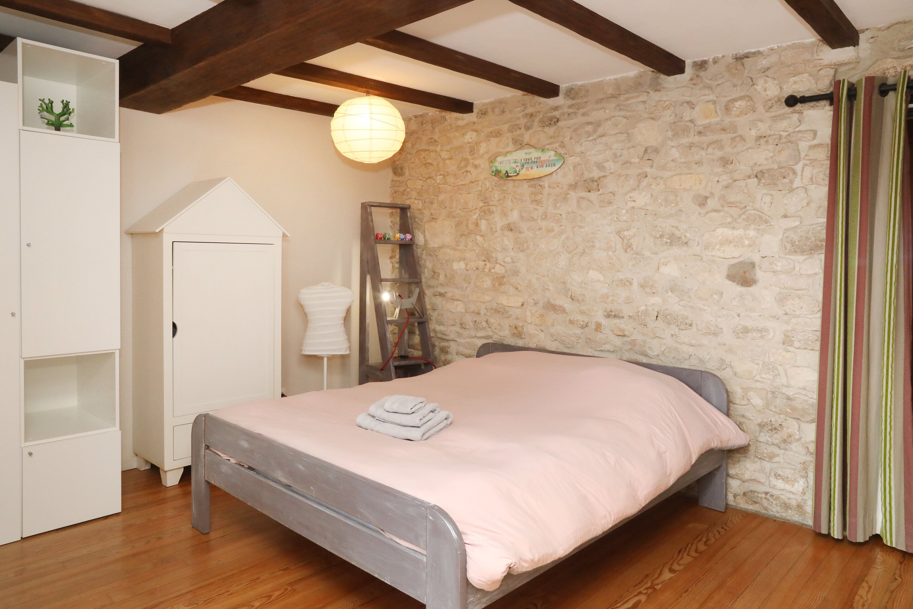 A bedroom with a stone wall, a bed with a pink bedspread, a wardrobe, and a small shelf with decorative items.
