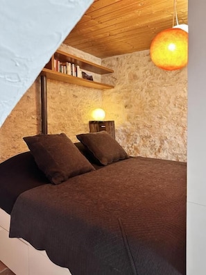Un dormitorio con pared de piedra, techo de madera, cama con ropa oscura, repisa con libros y una lámpara colgante.