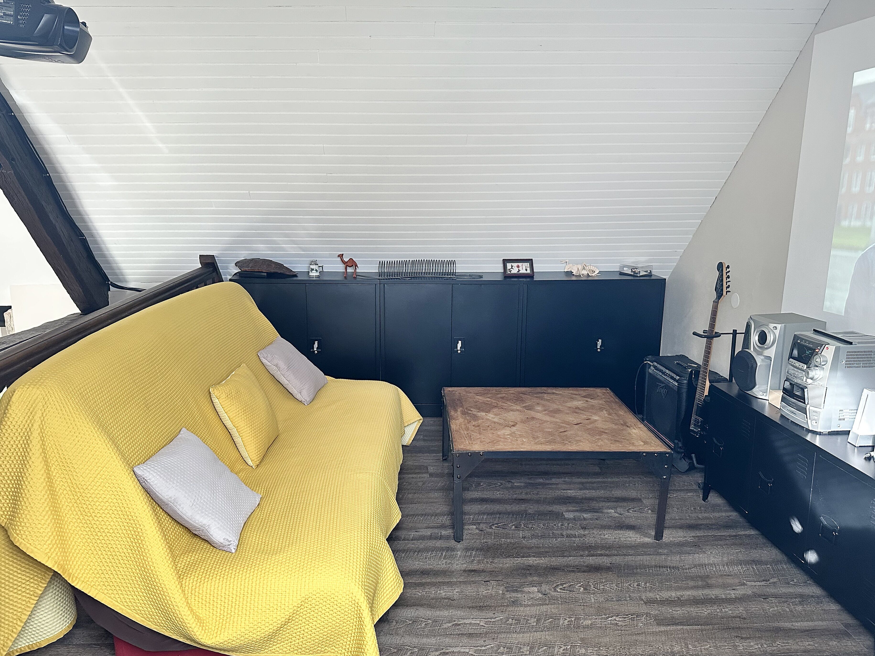 Una habitación con un sofá amarillo, una pequeña mesa de madera, un mueble negro con varios objetos y una guitarra.