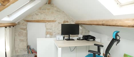 Una habitación con techo inclinado, un escritorio con computadora, una silla y una estantería.