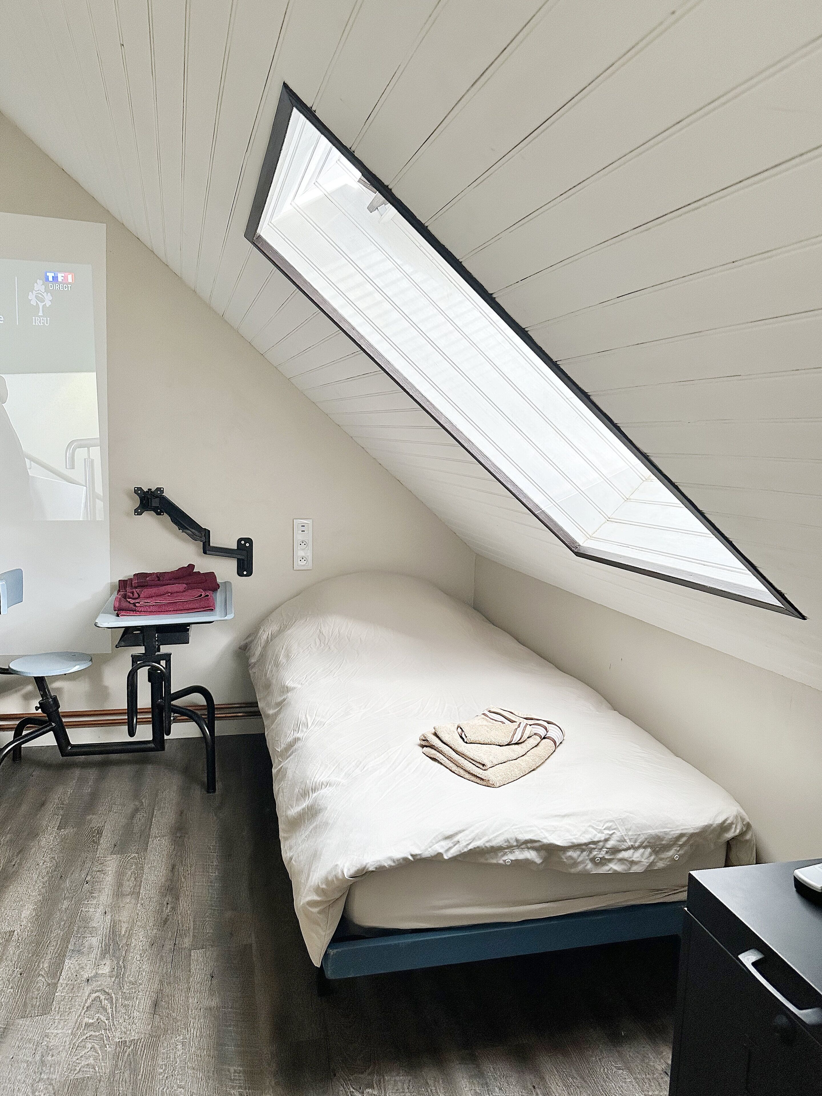 Ein kleines Zimmer mit einem Einzelbett, schrägem Dach, Schreibtisch und Stuhl sowie einem Heizkörper.