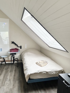 A small, single-bed room with a slanted ceiling, a desk with a chair, and a radiator.