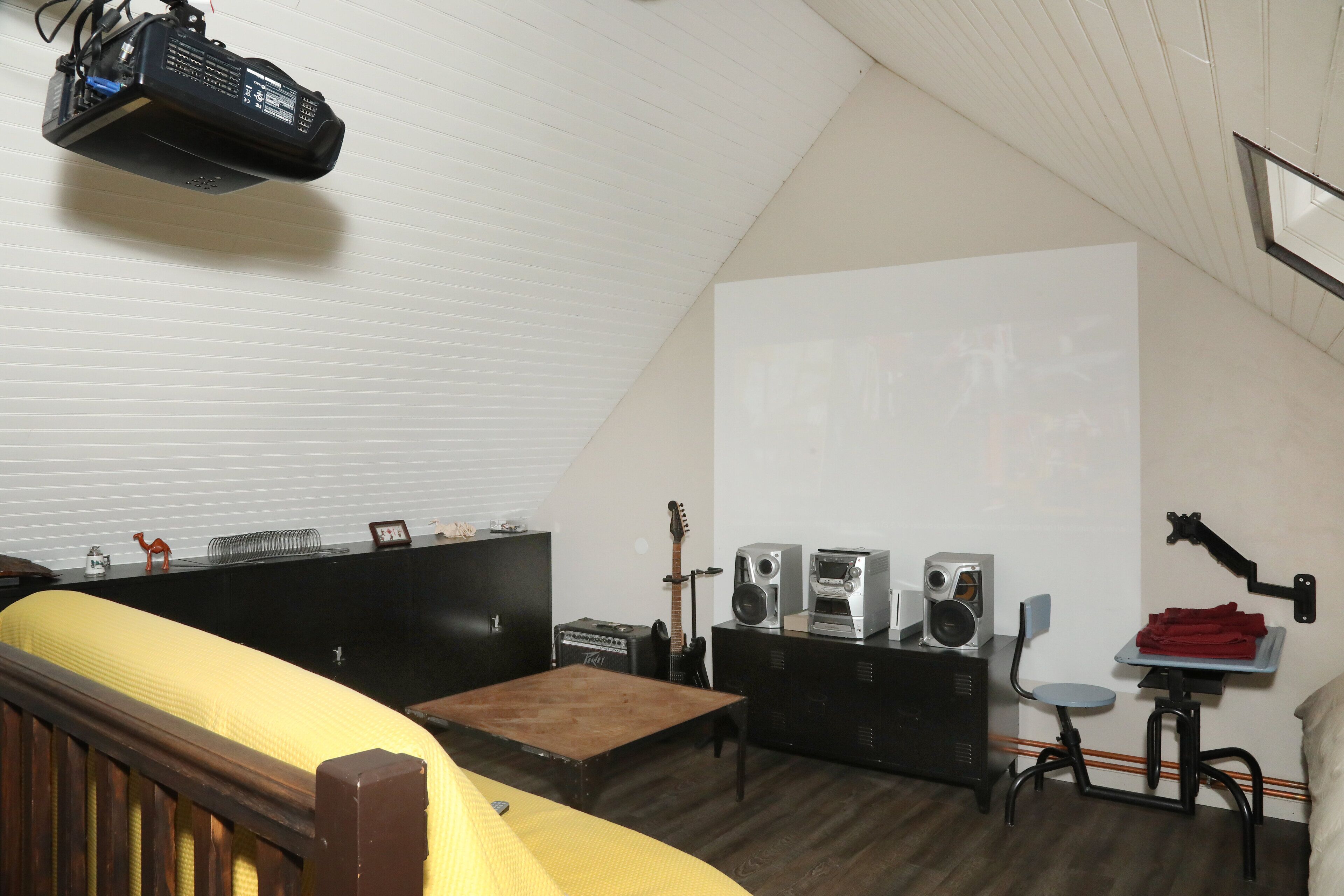 Una habitación con una cama, una mesita, una guitarra, altavoces y un proyector.