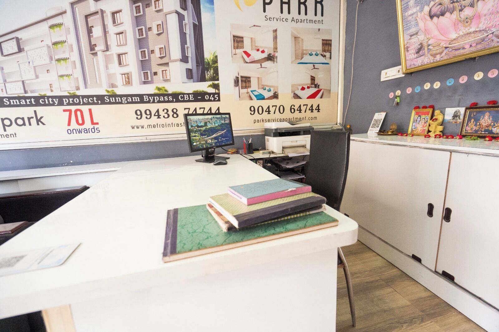 A reception desk with a computer monitor, a telephone, and a stack of books. There is a large poster with text and images of apartments.