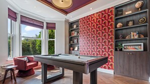 A billiards table in a room with a view of greenery, a red patterned wall, and a bookshelf with various items. - Luxury 8 Bed House, Games Room & Free Parking (Bedford)