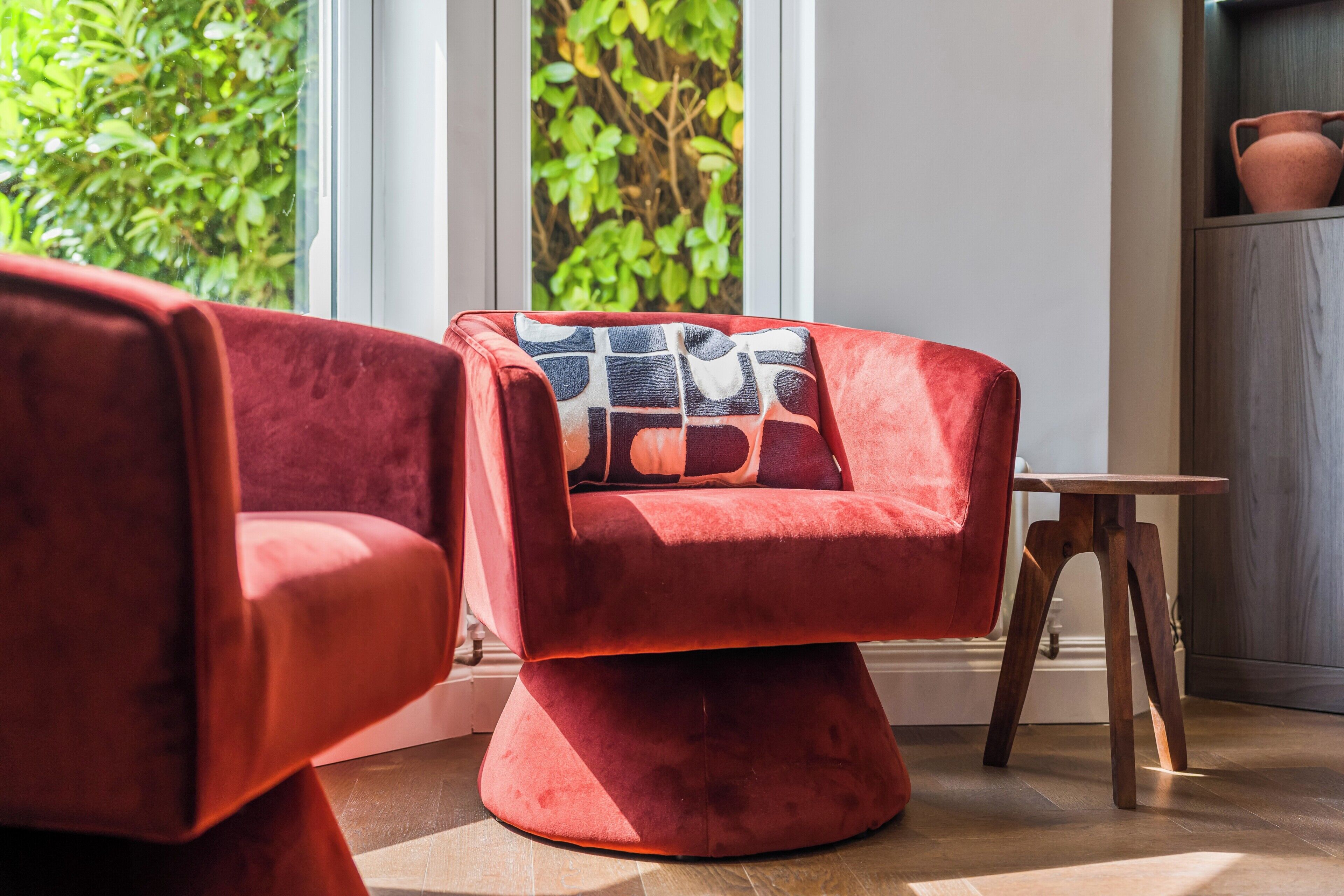 A modern living room with a red armchair, a wooden side table, and a view of greenery through a window.
