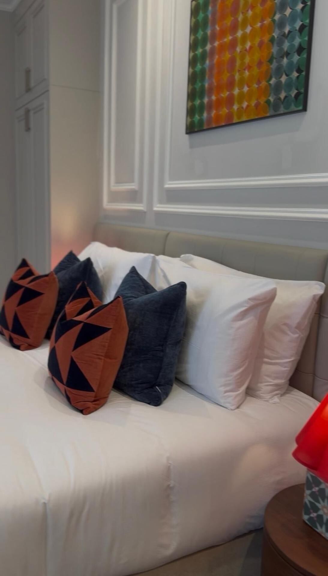 A bed with white bedding and multiple pillows, a colorful abstract painting on the wall, and a small round table with a red object on it.