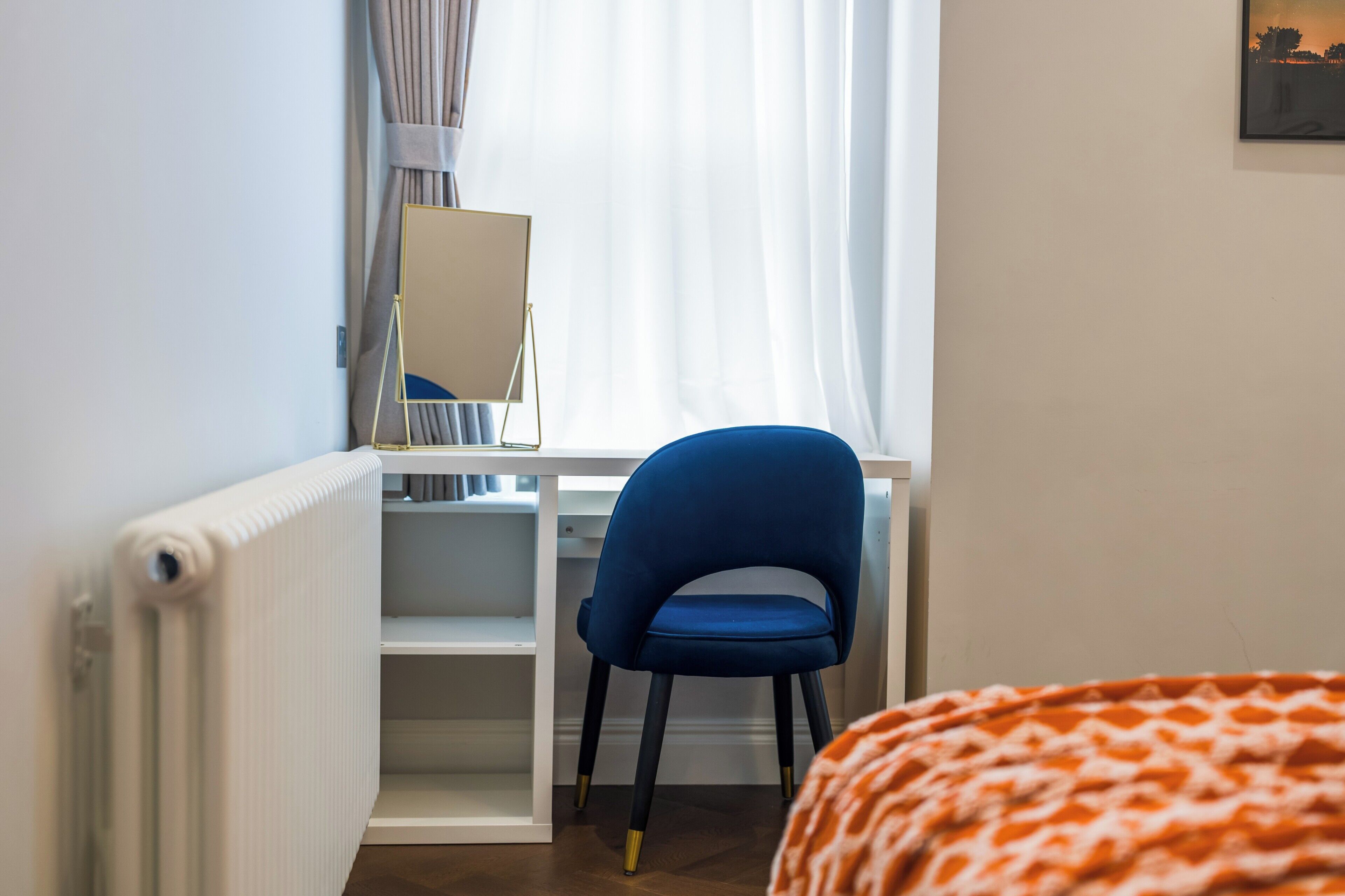 A bedroom with a bed, a desk with a chair, a radiator, and a window with curtains.