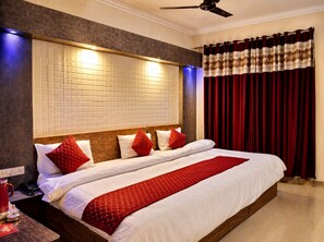 Quarto de hotel com uma cama grande, cabeceira iluminada, ventilador de teto e cortinas vermelhas.