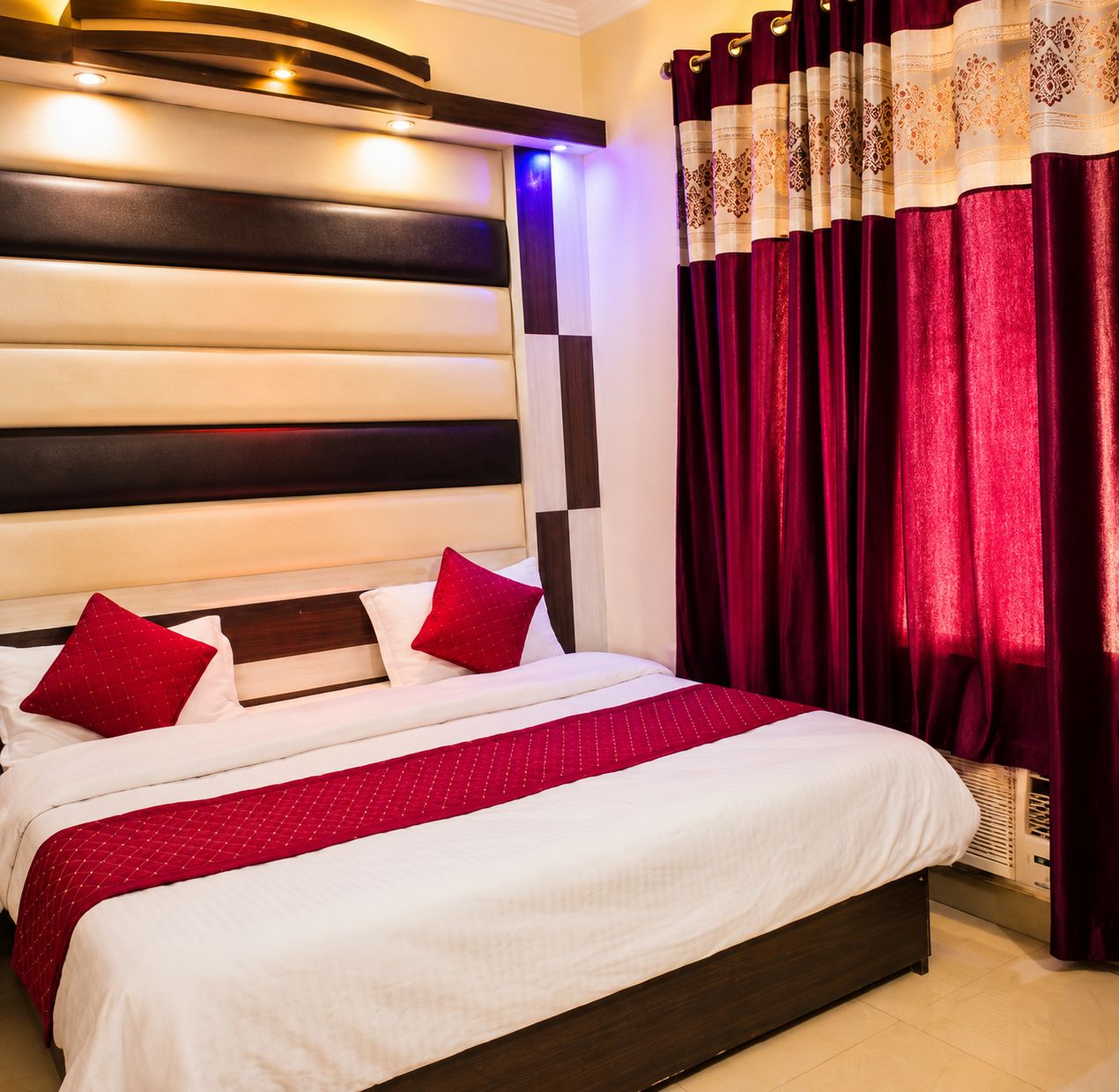 A hotel room with a large bed, red and white bedding, a dark headboard, and red curtains.