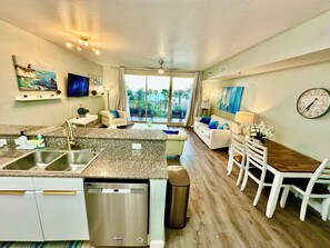A modern kitchen with a dishwasher, a dining area with a white table and chairs, a sofa with blue cushions, and a large window offering a view of palm trees. - 3BR 3BA Beachfront 8PPL.Most gorgeous resort- 3rd floor unit and 3rd floor parkg (Panama City Beach)