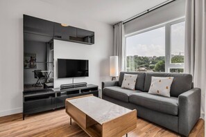 A modern living room with a flat-screen TV, grey sofa, wooden coffee table, and a city view through large windows.